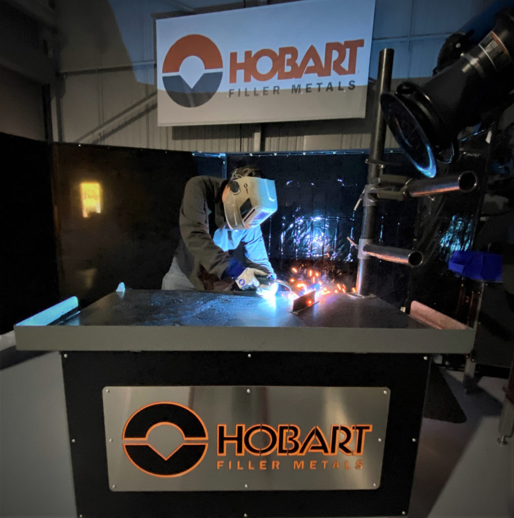 Welder MIG welding a thin weld coupon on a table with Hobart banner behind him